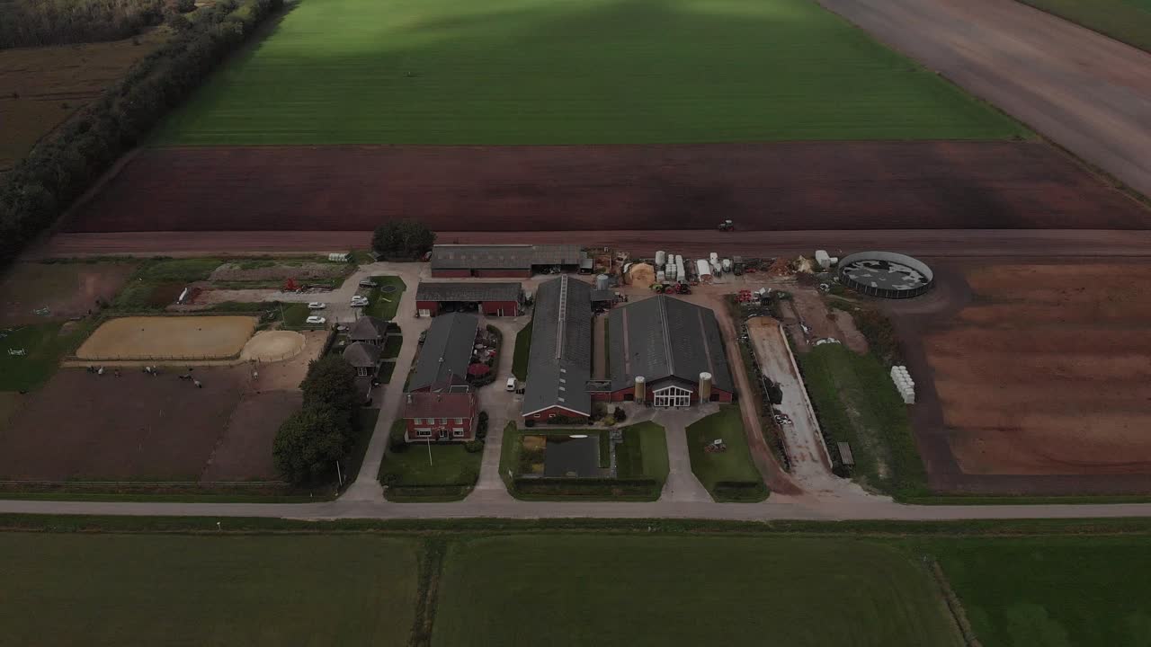 descenso e inclinación aéreos que muestran una granja agrícola holandesa con la luz del sol golpeando los parches de prado y tierras de cultivo alrededor de las construcciones relacionadas con el negocio