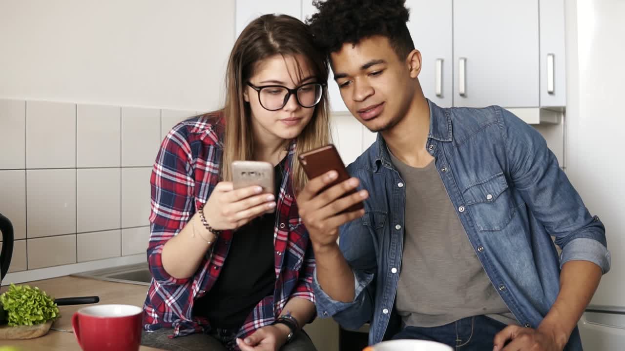 Attractive guy and a girl both in their 20's scrolling their smartphones, exchanging information while sitting in a comfy spacious kitchen.