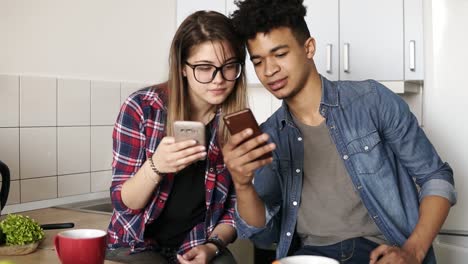 Attractive-guy-and-a-girl-both-in-their-20's-scrolling-their-smartphones,-exchanging-information-while-sitting-in-a-comfy-spacious-kitchen.