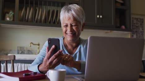 Glückliche-ältere-Frau-Mit-Gemischter-Abstammung,-Die-Smartphone-Und-Laptop-In-Der-Küche-Benutzt