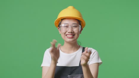 close up of asian woman worker wearing goggles and safety helmet smiling and clapping her hands while standing in the green screen background studio