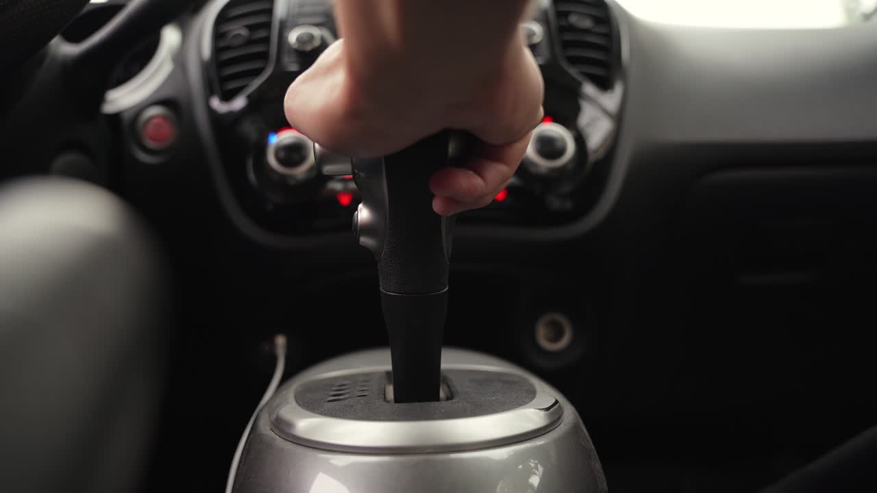 La mano di una persona su un cambio automatico all'interno di un'auto moderna. La luce naturale illumina il cruscotto e la console centrale durante un tragitto urbano o un viaggio su strada.