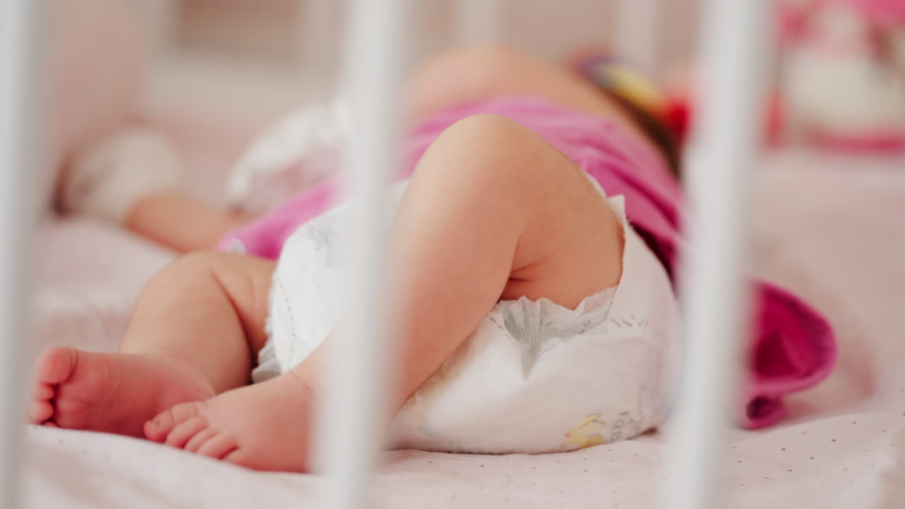 Close up of a baby's legs and diaper as the infant rests in a crib, seen through the crib bars
