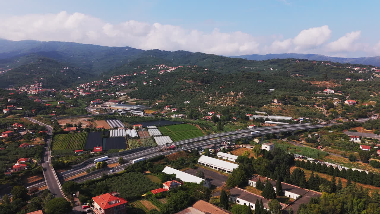 High drone shot moving forward toward a road cutting through residential areas, with visible traffic, trees, and surrounding mountains near Diano Marina