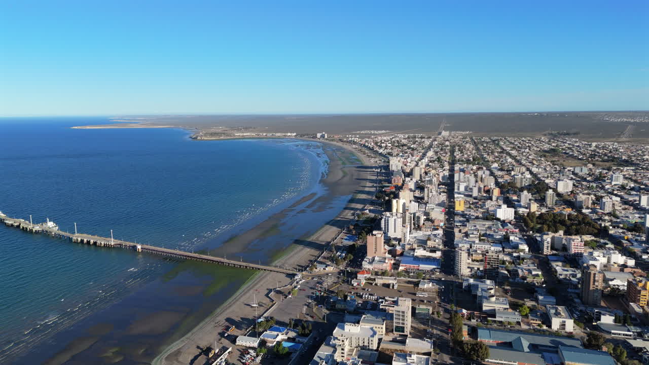 Drone shot over Puerto Madryn’s shoreline, with ocean, dock, city buildings, and a clear blue sky. Distant horizon in background. Shot on 4K at 60fps.