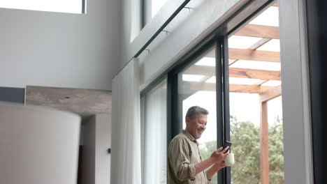 hombre biracial mayor feliz usando teléfono inteligente por la ventana en la sala de estar, espacio de copia, cámara lenta