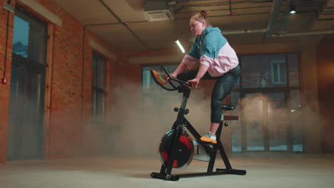 Mujer-Atlética-Montando-Una-Rutina-De-Entrenamiento-En-Bicicleta-Estacionaria-Giratoria-En-Un-Gimnasio-De-Niebla,-Pérdida-De-Peso-En-Interiores