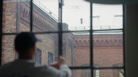 person gazes thoughtfully through a snow-dappled window, historic brick building backdrop