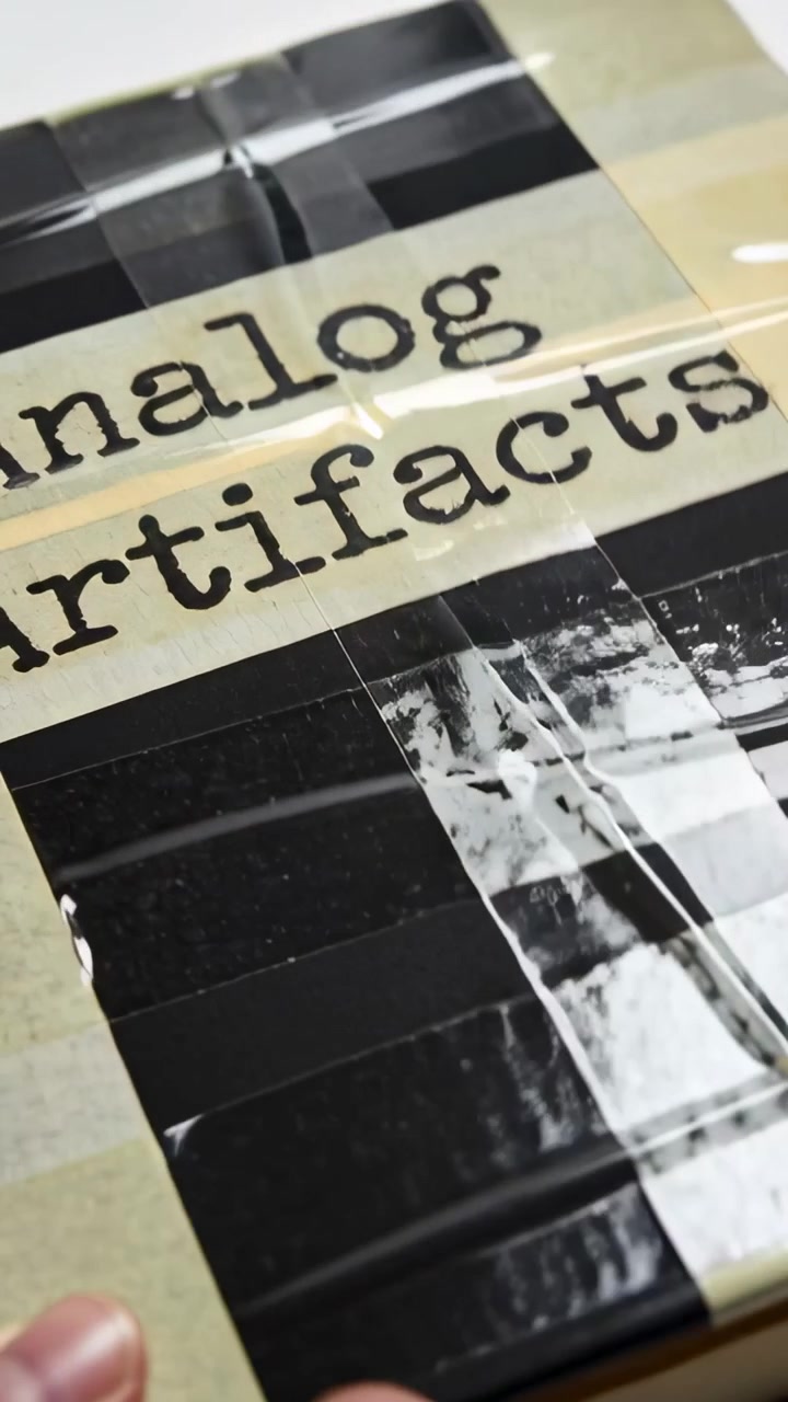Lodret video: Panorerer over indpakket bog og analoge genstande på skrivebord, hånd justerer tape for at afsløre etiket.