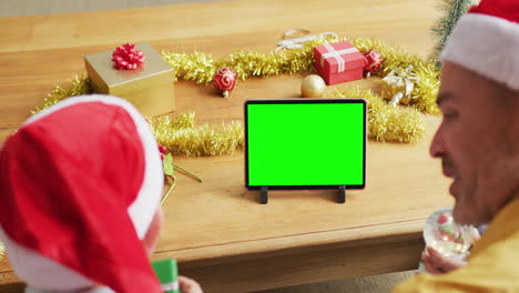 padre y hijo caucásicos felices en sombreros de santa haciendo una llamada de video de navidad de tableta, con pantalla verde