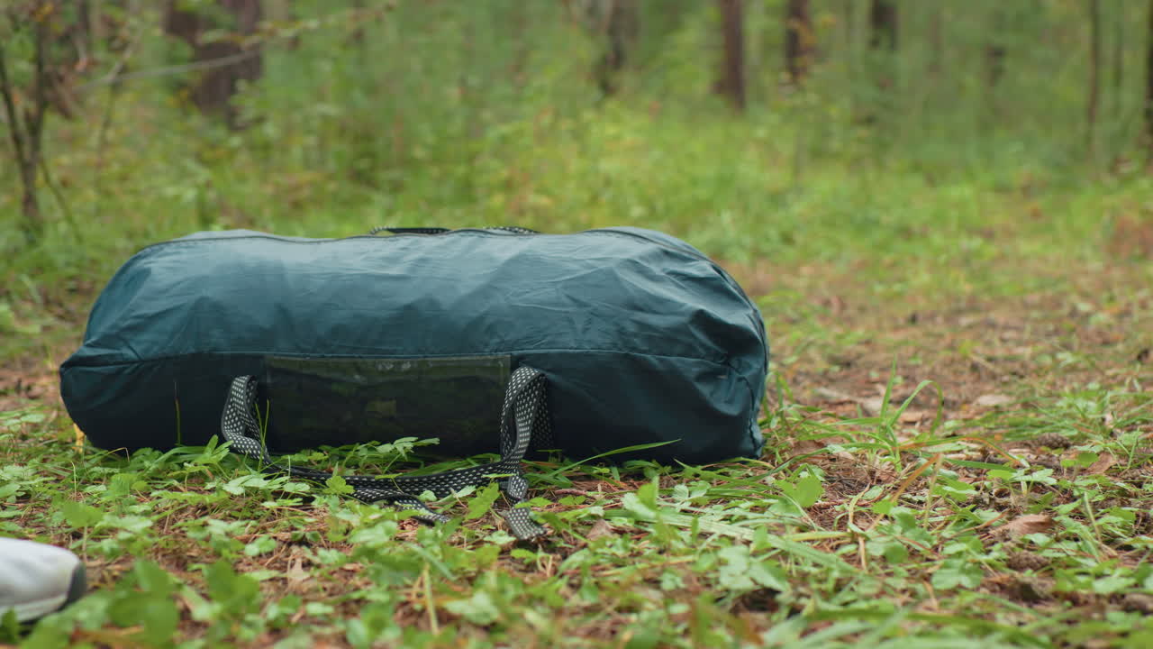 close up of green duffel bag being dropped onto forest ground surrounded by leaves, pine needles, and grass, showing beginning of rest or camping moment in outdoor woodland environment