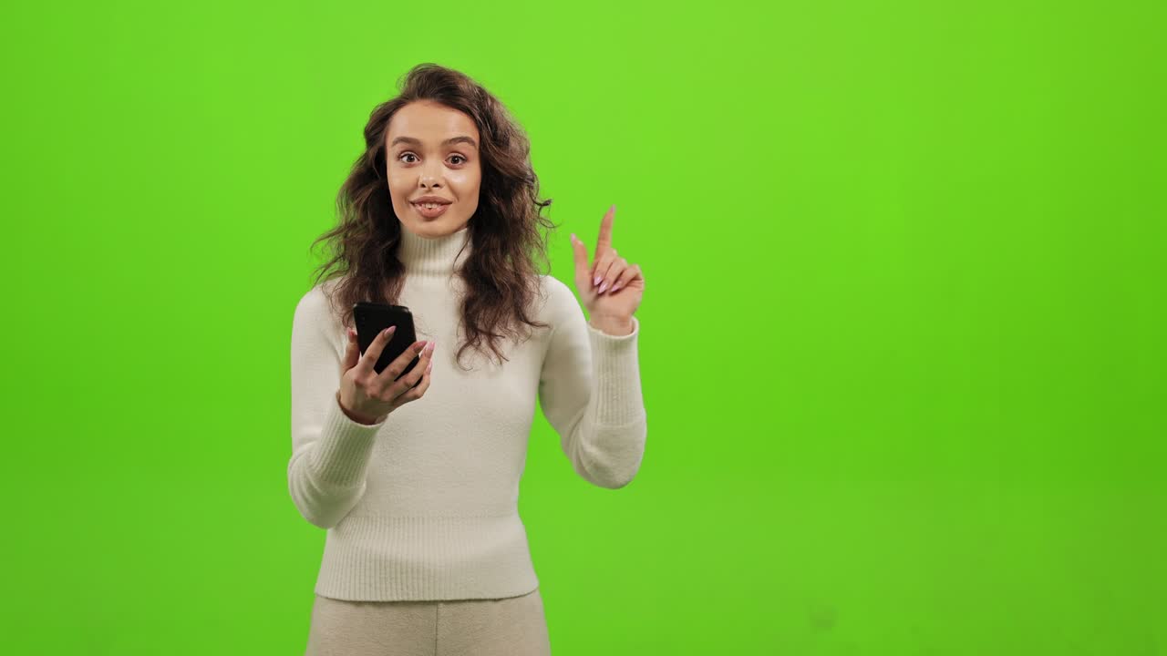 The woman is looking through social networks on her smartphone. She is raising her head and looking at the camera. She has an idea and raises her finger up. She is standing on a green background. Green screen. 4K.
