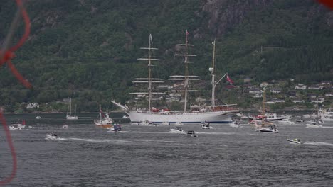 a tall sailing ship with a norwegian flag sails through a busy fjord surrounded by boats