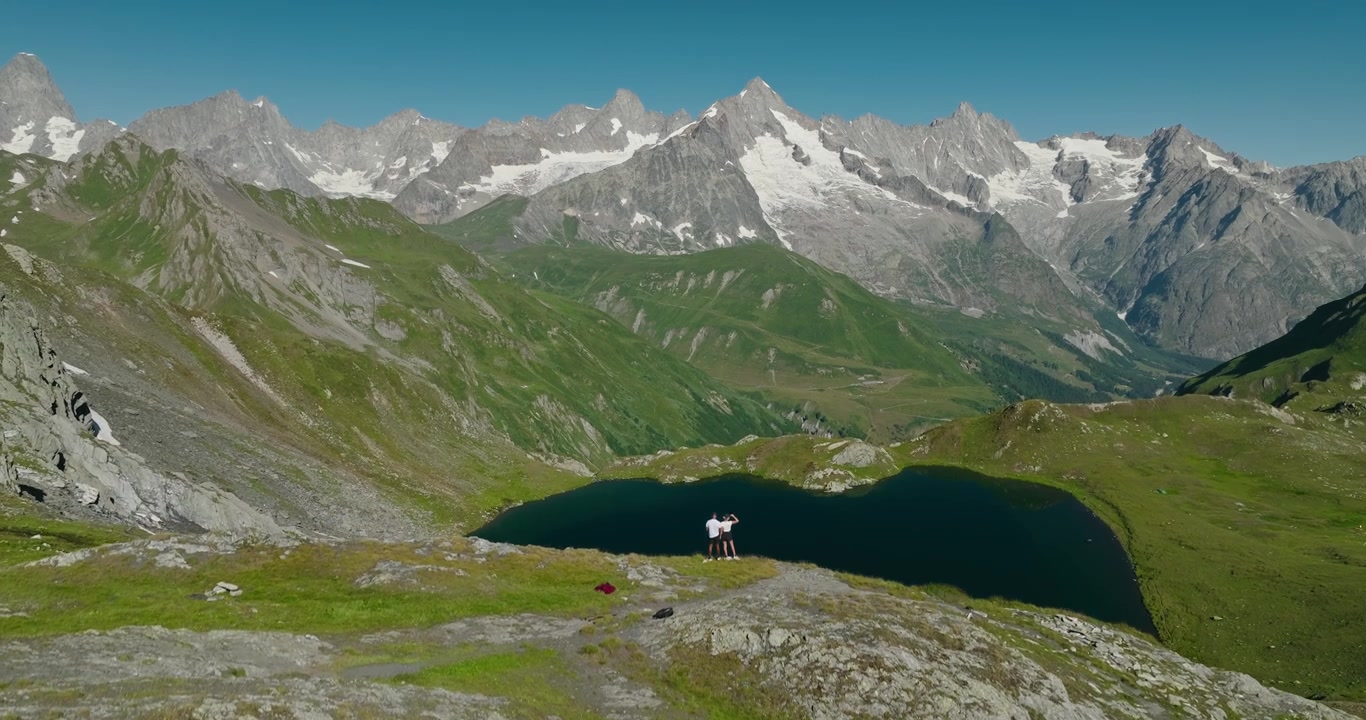 Un drone si alza da dietro una cresta montuosa per rivelare due escursionisti in piedi su una scogliera sopra un lago alpino, con ghiacciai distanti e imponenti vette svizzere sotto un cielo estivo limpido e azzurro
