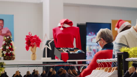 Una-Pareja-De-Ancianos-Feliz-Después-De-Encontrar-El-Regalo-De-Navidad-Perfecto-Para-Su-Hijo-Mientras-Compraba-En-Una-Tienda-De-Ropa-Decorada-Con-Navidad.-Clientes-Navegando-Por-La-Ropa-Durante-La-Temporada-De-Vacaciones-De-Invierno.