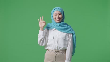 아시아 여성이 미소 짓고 녹색 스크린 배경 스튜디오에 서있는 동안 카메라에 은 몸짓을 보여줍니다.