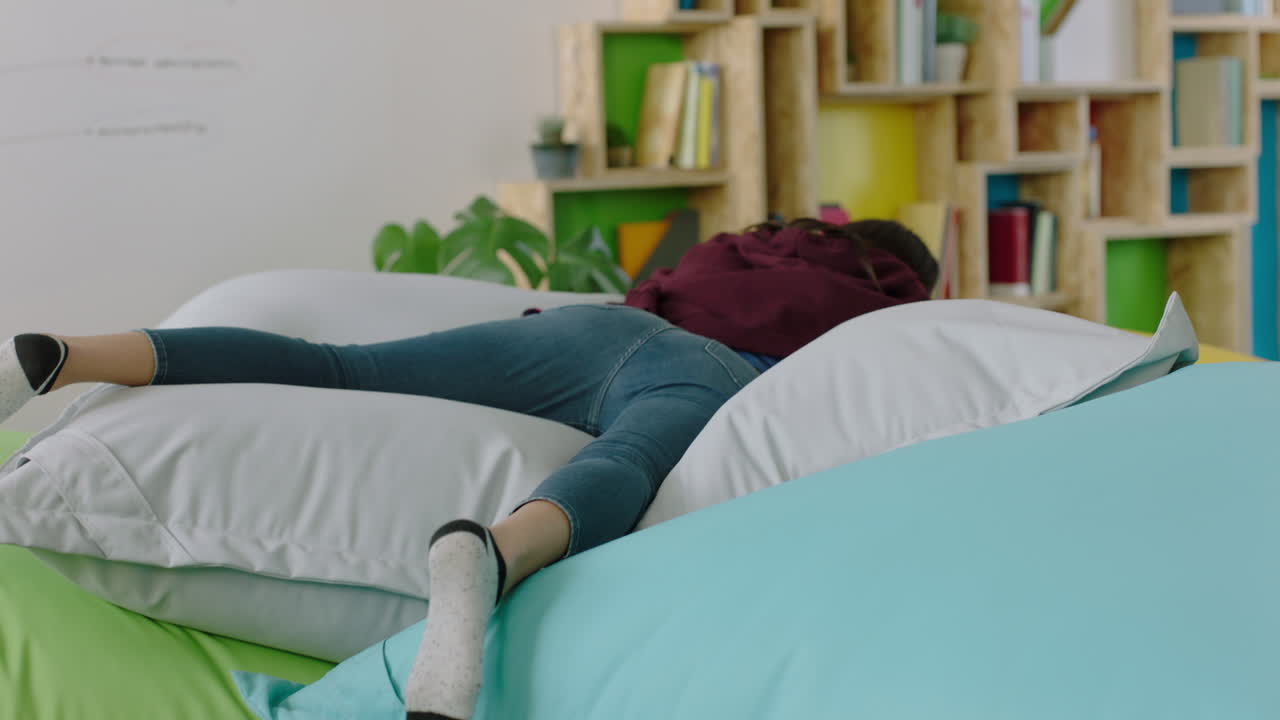 행복한 백인 여성 학생이 다채로운 베개에 뛰어들고 여자가 휴식을 취하고 트렌디한 사무실 작업장에서 점심 휴식을 즐기고 있습니다.