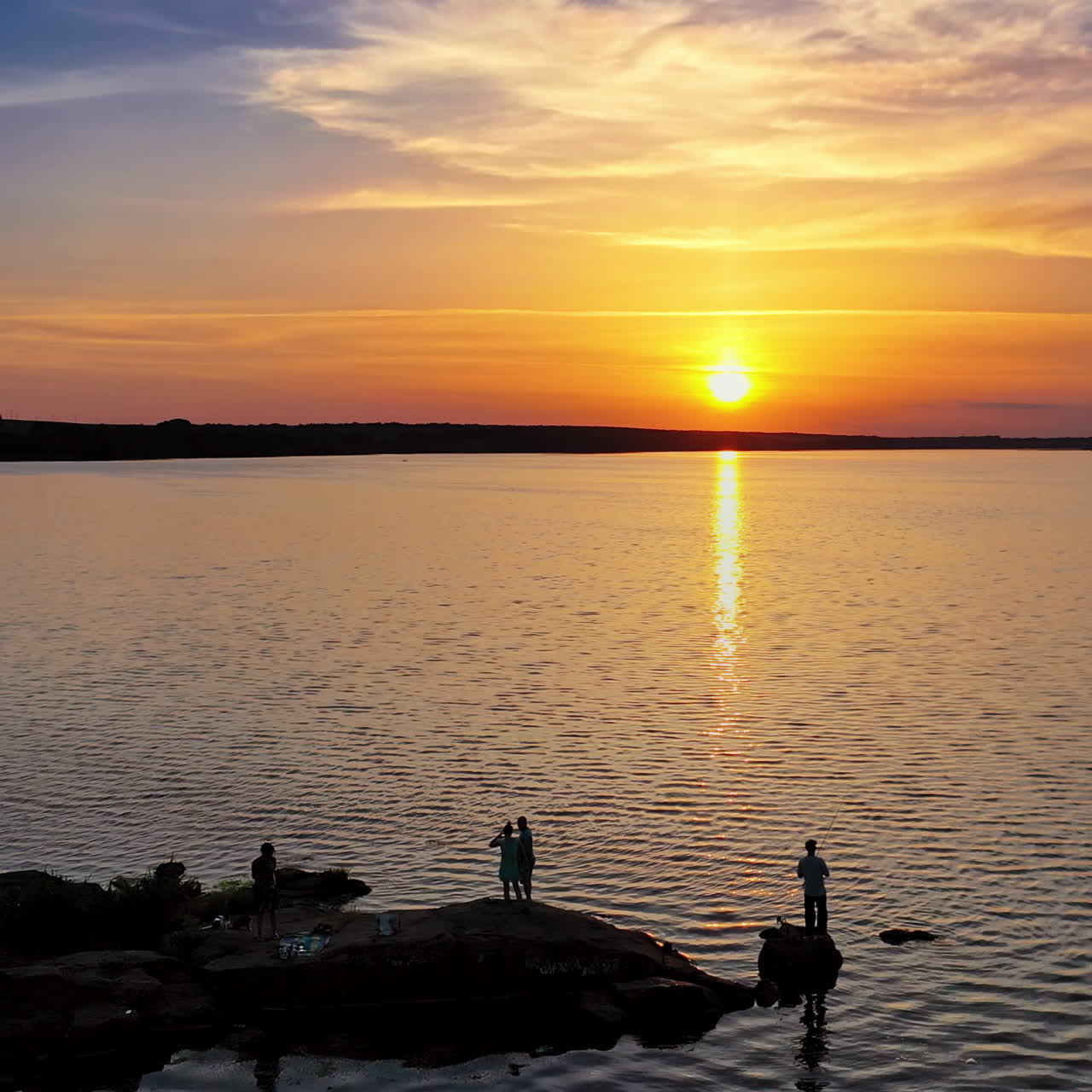 View from a drone on the river in the evening. People stand on long stones situated in water at sunset. Camera moves forward.