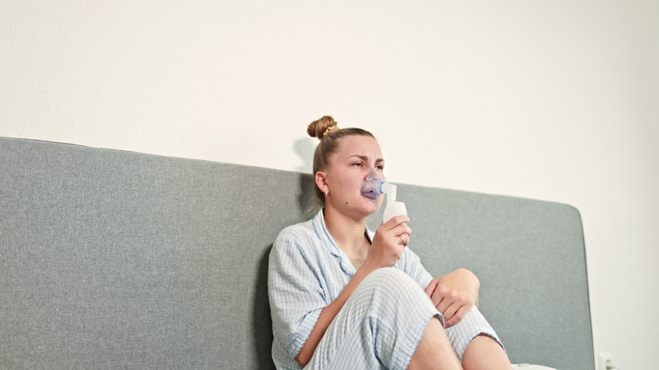 Jeune femme assise sur le lit, mettant un masque sur son visage pour une inhalation thérapeutique. Femme faisant une inhalation pour traiter la toux. Traitement par nébulisation à domicile. Auto-traitement avec une solution saline et un inhalateur.