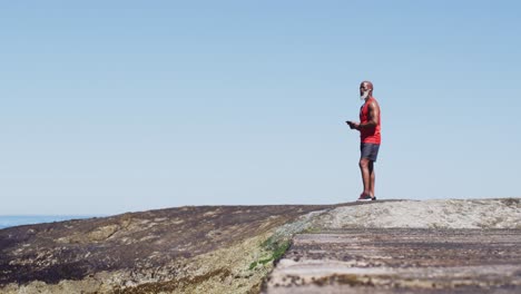 hombre afroamericano de alto nivel haciendo ejercicio usando un teléfono inteligente en rocas junto al mar