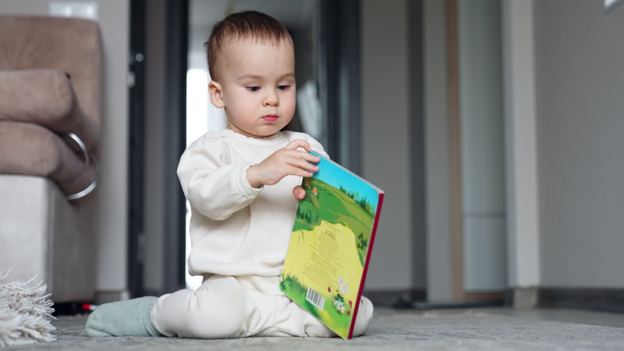 Little baby boy focused on the book in his hands. Lovely kid sits on the floor trying to open the book.