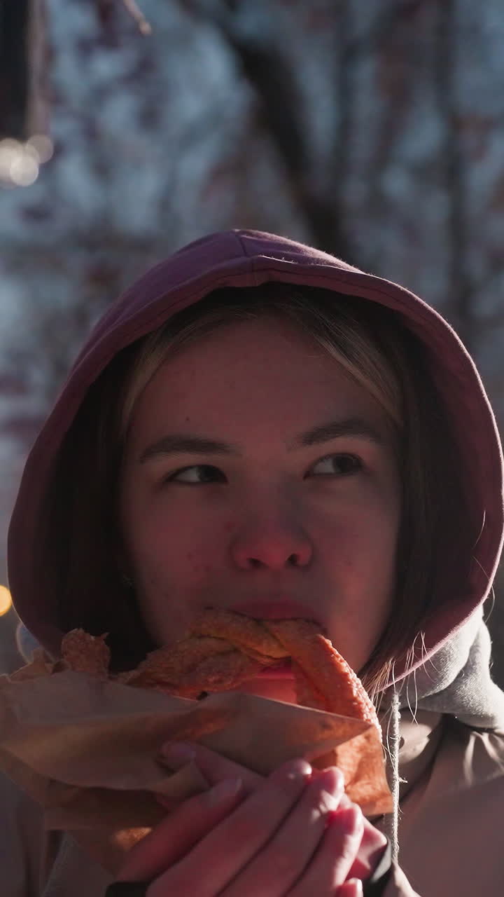 mujer joven de pie al aire libre al anochecer en un parque nevado, colocando suavemente comida en la casa de los pájaros mientras sostiene la comida en la mano, rodeada de tierra cubierta de nieve, luces de la calle suaves en el fondo