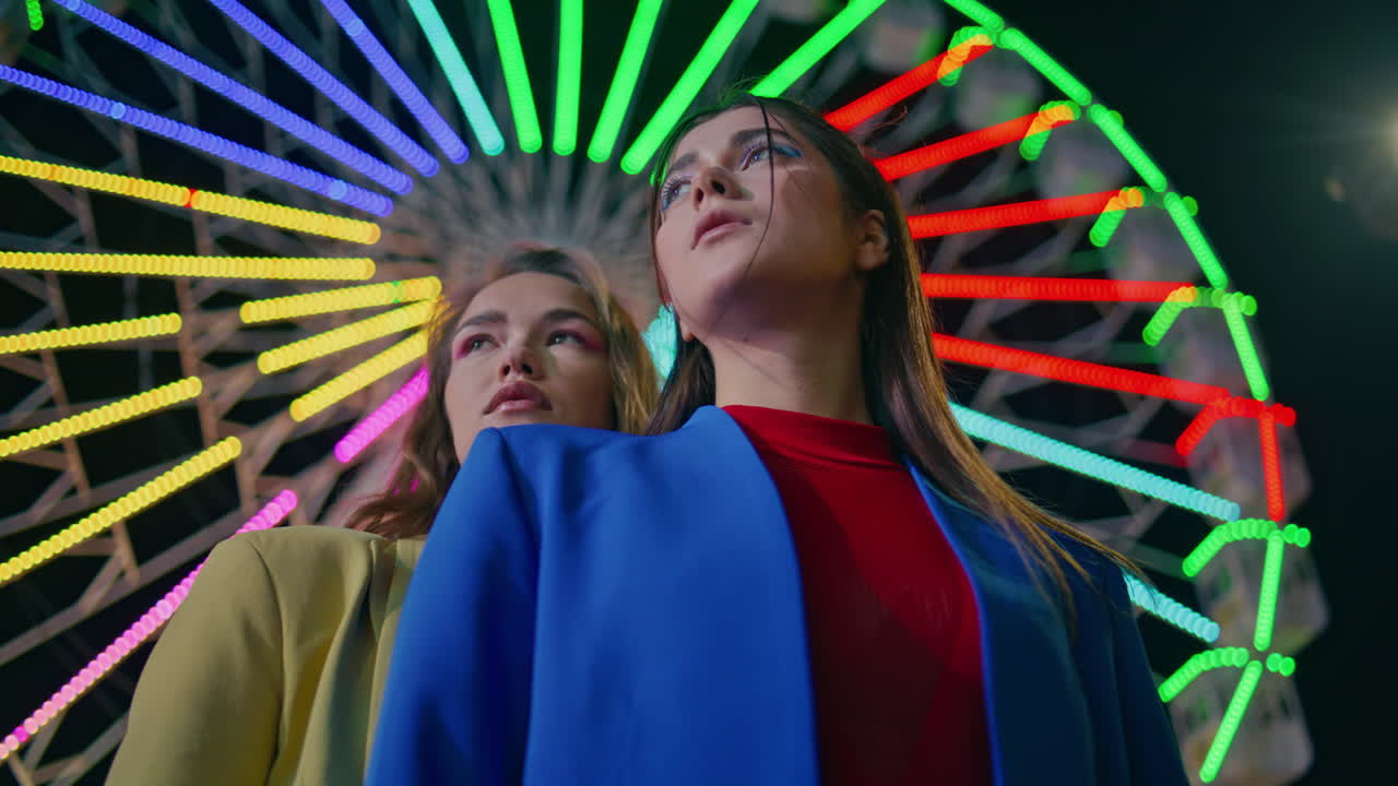 Closeup girls rest ferris wheel at night. Two fashion models pose at illuminated