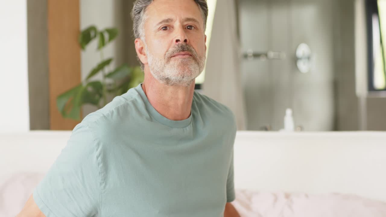 Uomo adulto maturo seduto sul letto che si volta verso il bagno, sistemandosi la camicia mentre la telecamera si allontana.