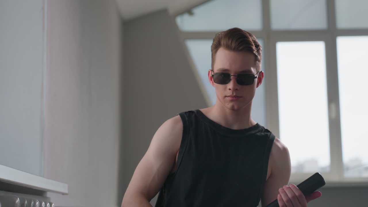 Youthful security guard vibe with neck tattoo and black tank top stands by loft window, wearing dark sunglasses and gripping sleek security baton, sunlight casting edgy shadows on confident face
