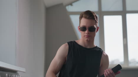 youthful security guard vibe with neck tattoo and black tank top stands by loft window, wearing dark sunglasses and gripping sleek security baton, sunlight casting edgy shadows on confident face