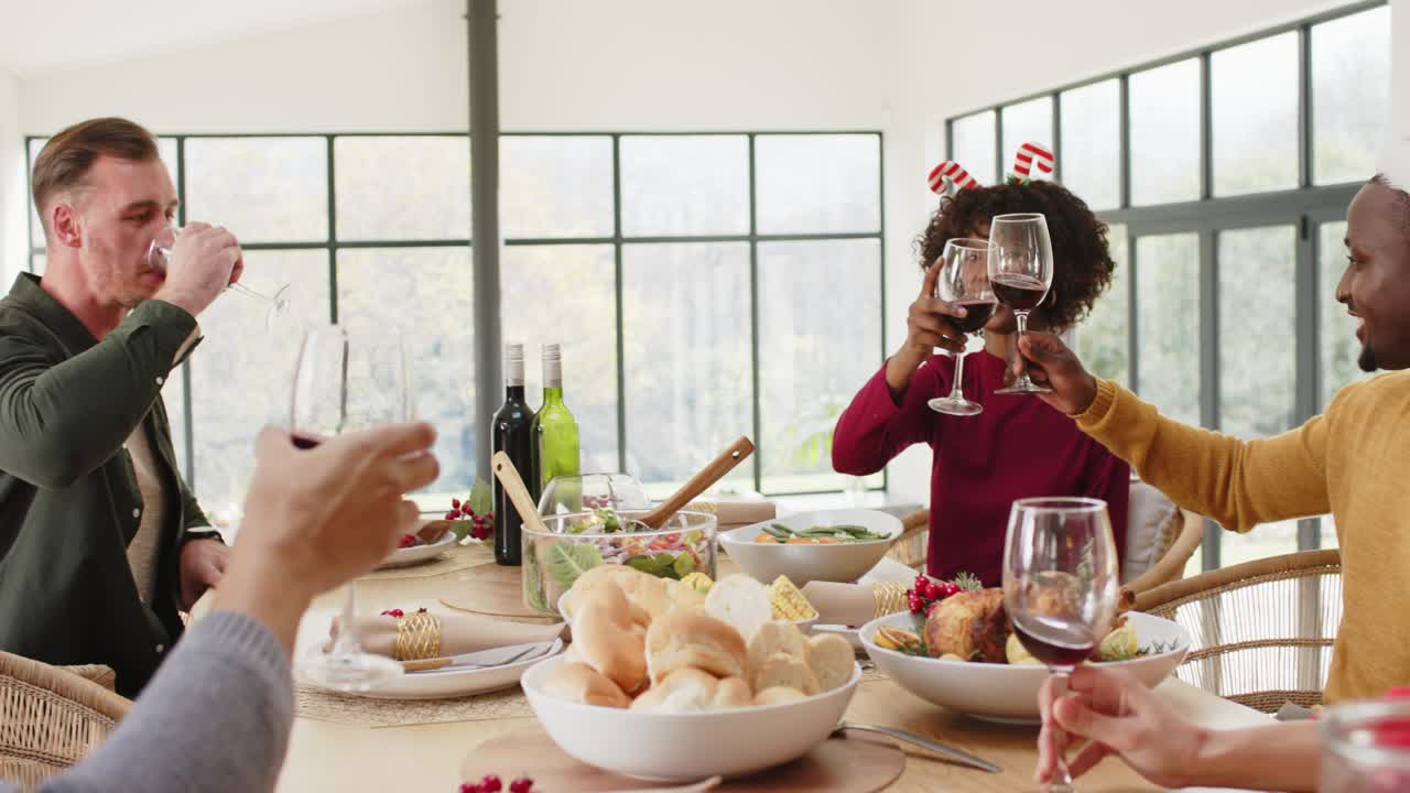Amis levant des verres de vin rouge à une table en chêne, trinquant et sirotant du vin pour un repas de fête.