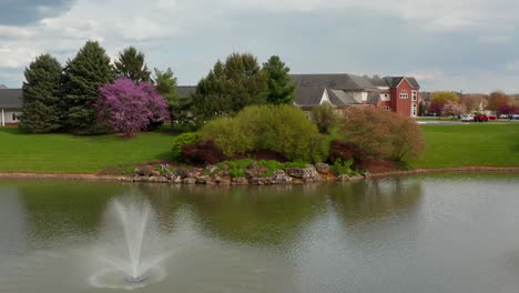 Fountain-at-pond,-aerial-rises-to-reveal-medical-retirement-home-building-in-USA