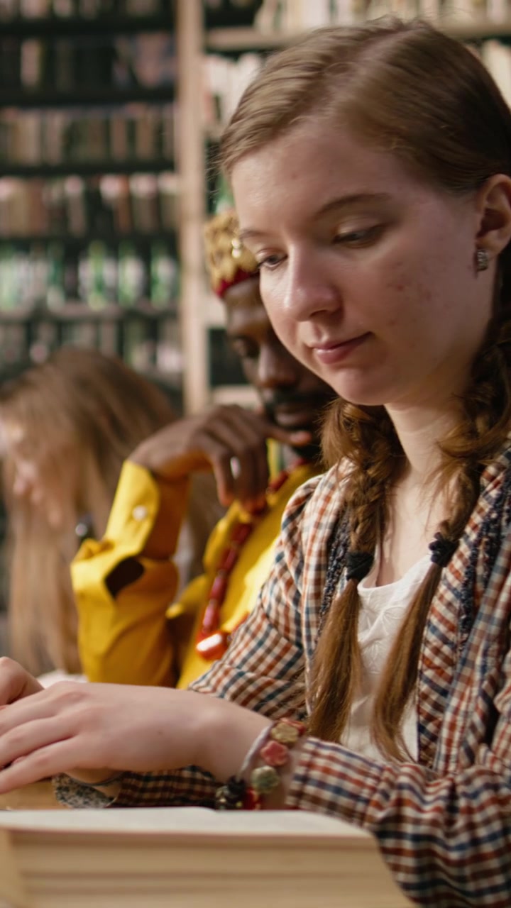 Video verticale: Studentessa che studia a un tavolo condiviso di biblioteca con laptop e libro aperto
