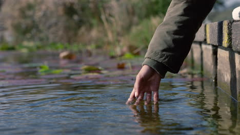 Cerrar-La-Mano-De-Una-Mujer-Salpicando-Agua-Disfrutando-De-Tocar-Un-Estanque-Fresco-En-El-Parque-Natural-Al-Aire-Libre