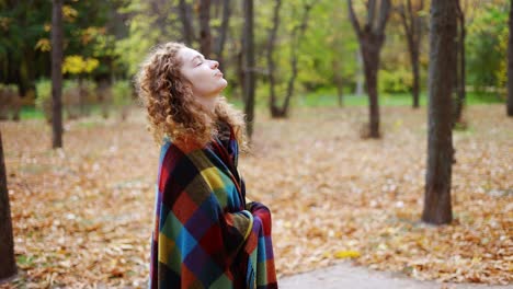 young woman standing in golden autumn park or forest covering herself in plaid enjoying good day weather