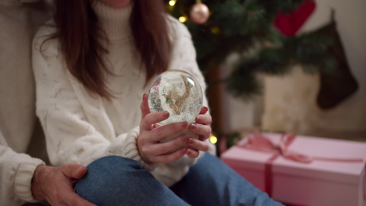 fotografía en primer plano de una chica morena con un suéter blanco sacudiendo una esfera de vidrio de año nuevo cerca de su novio en una habitación acogedora decorada para el año nuevo en invierno