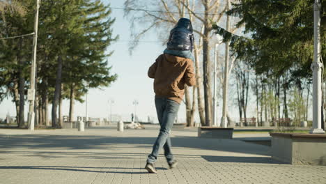 un padre corre juguetón por un camino pavimentado bordeado de árboles mientras lleva a su hijo en los hombros, el padre sostiene las piernas del niño para garantizar el equilibrio, ambos vestidos con chaquetas