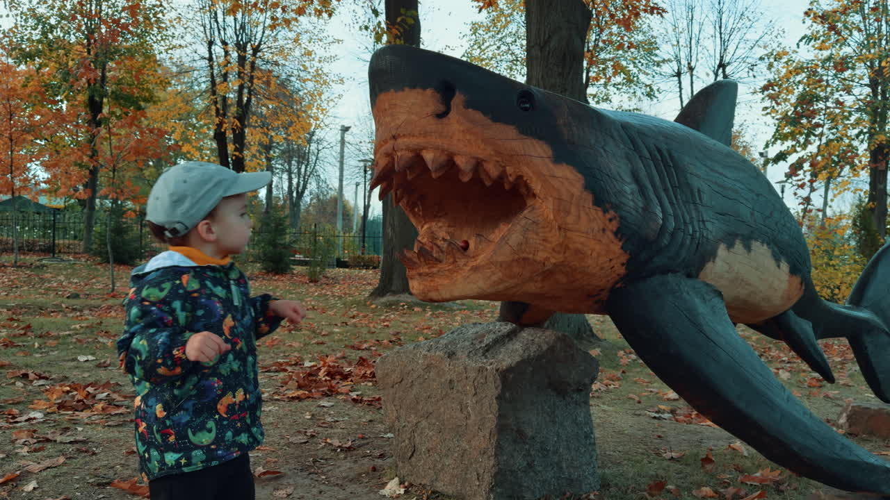 Baby boy throwing the chestnuts into the mouth of a wooden shark sculpture. Toddler playing outdoors in autumn.
