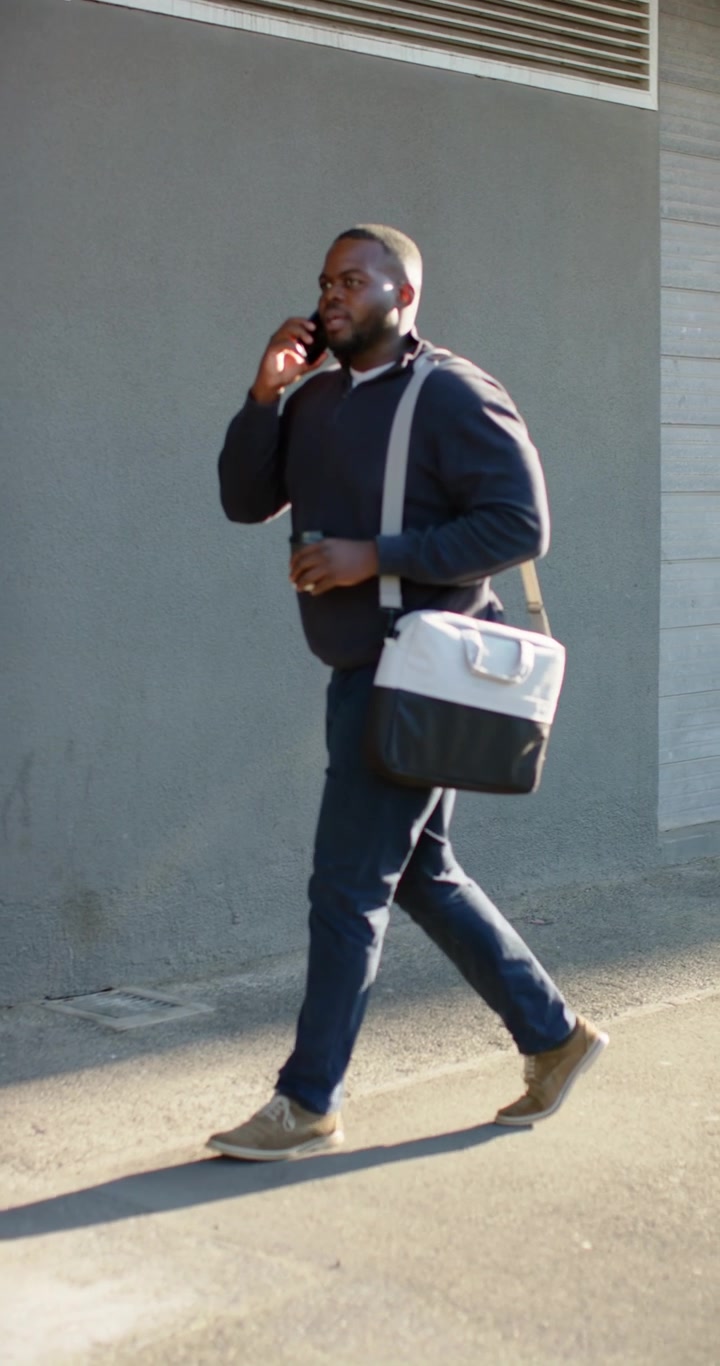 Verticale video: Afro-Amerikaanse man komt in beeld, loopt op trottoir, gebruikt smartphone met tas