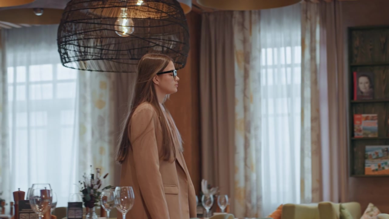 Donna che cammina accanto a un tavolo apparecchiato con bicchieri e decorazioni, luci a sospensione che proiettano un bagliore cinematografico, la fotocamera cattura primi piani di bicchieri da vino riflettenti, profondità di campo ridotta, scaffali e tende sfocati.