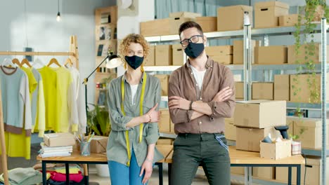 young caucasian man and woman in mask looking at camera in good mood sitting on table in own shop with clothes