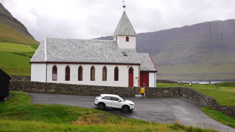 man entering yard of white vidareidi church on meadow in mountains, faroe islands