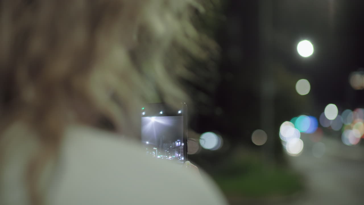Rear view of female photographer taking photo of urban street with smartphone at night, capturing illuminated road with car passing by, colorful bokeh lights in background