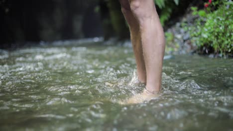 a male european hiking tourist is standing with bare feet in cold water creek a river close to a large waterfall cliff in the green forest jungle of tanzania