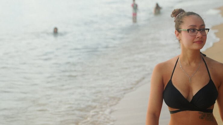 Primo piano di donna asiatica in riva al mare, che si sistema gli occhiali e lo chignon, collana delicata che cattura la morbida luce serale, onde leggere e sabbia bagnata ai piedi, sguardo riflessivo verso l'orizzonte, vlogger di viaggi