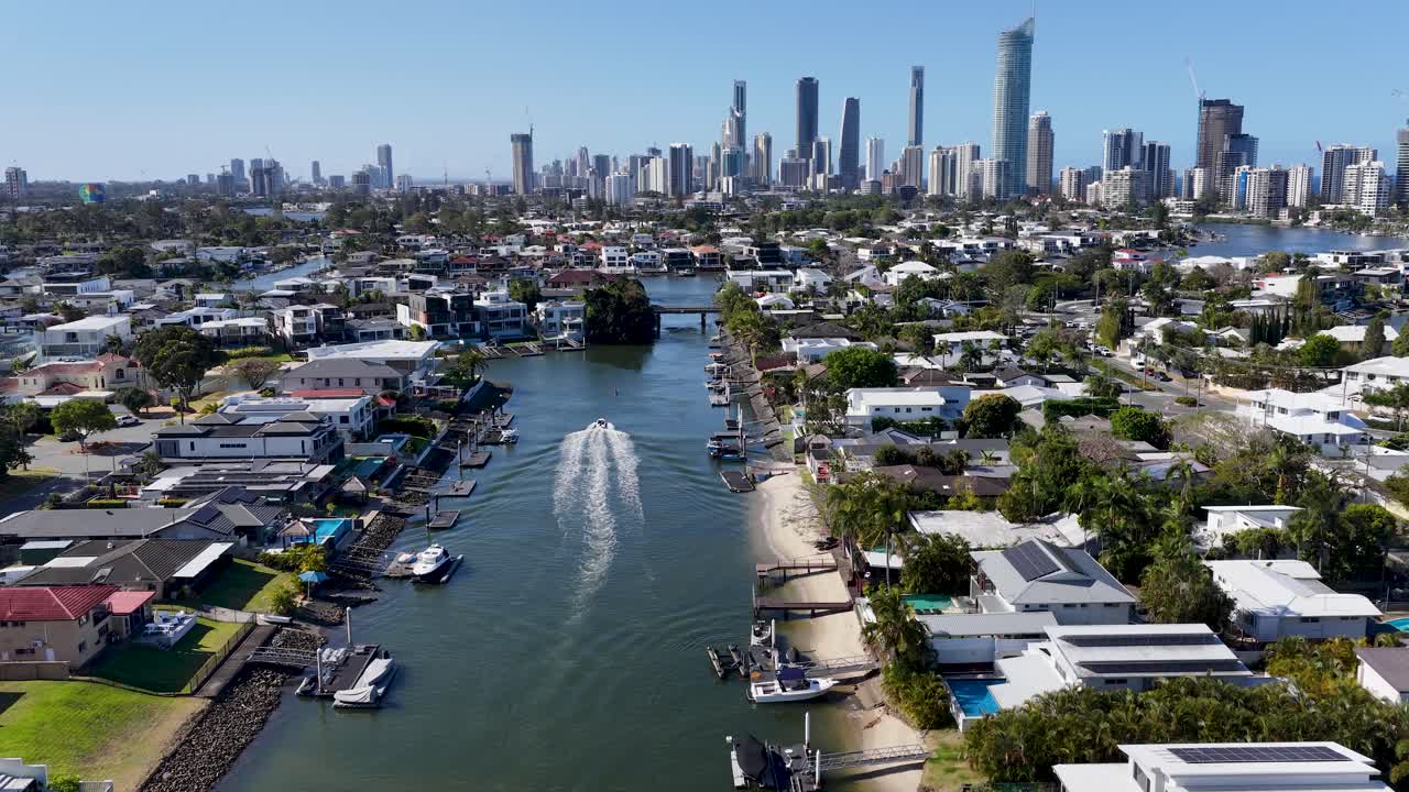 Drone follows motorboat along canal, modern houses, city skyline, bright daylight, smooth forward motion