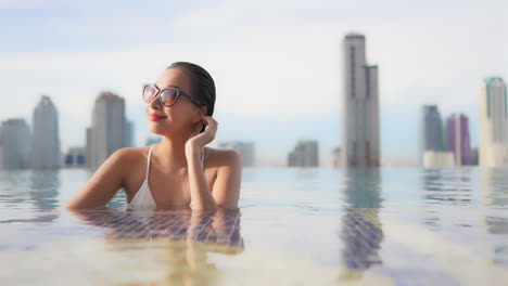 Mientras-Descansa-En-Una-Piscina-Infinita-En-La-Azotea-Respaldada-Por-El-Horizonte-Moderno-De-La-Ciudad-De-Bangkok,-Tailandia,-Una-Hermosa-Mujer-Joven-Gira-Su-Rostro-Hacia-El-Sol