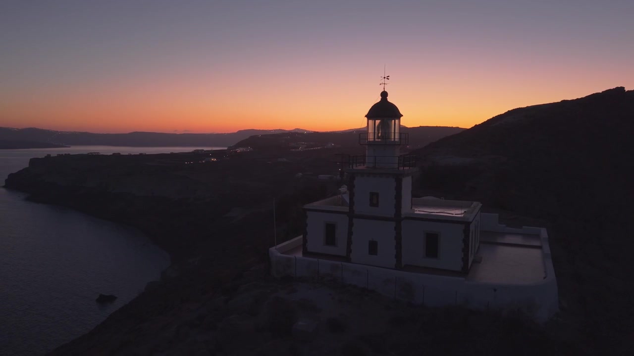 Oranžový horizont při východu slunce v Akrotiri na Santorini s ostrovním majákem s výhledem na Egejské moře a dramatické sopečné pobřeží.