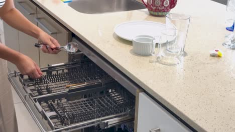 a woman loads plates, glasses, and cutlery into a built-in dishwasher in a bright, modern kitchen with overhead lighting and steady camera angles
