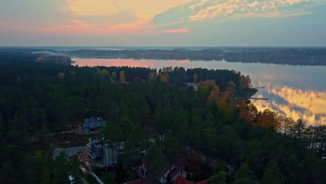 Aerial-drone-shot-over-wooden-houses-beside-a-large-lake-along-rural-countryside-surrounded-by-dense-green-vegetation-during-evening-time